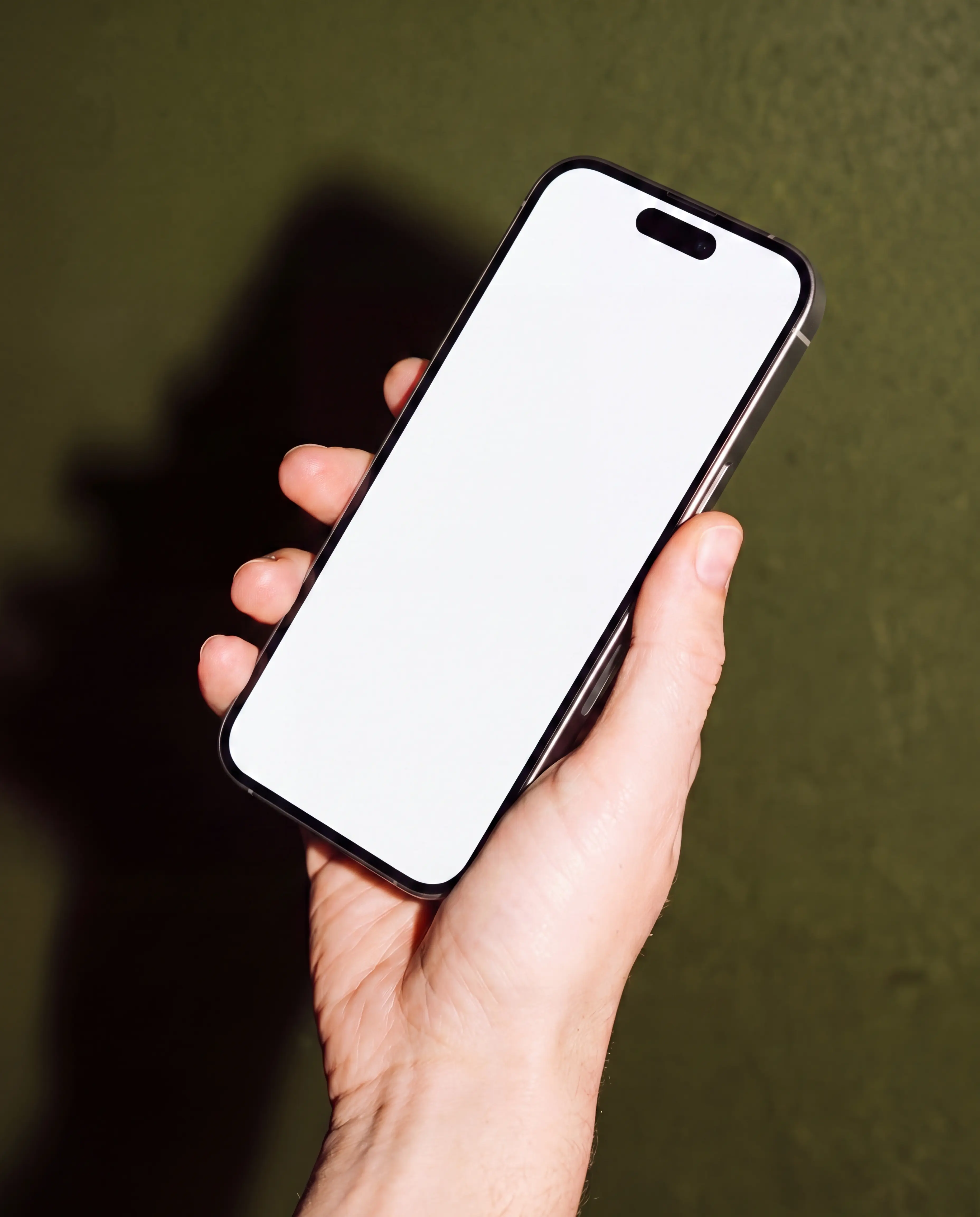 Close-up modern black iPhone held diagonally in one hand against a softly textured olive-green background, with an editable white vertical display and visible Dynamic Island