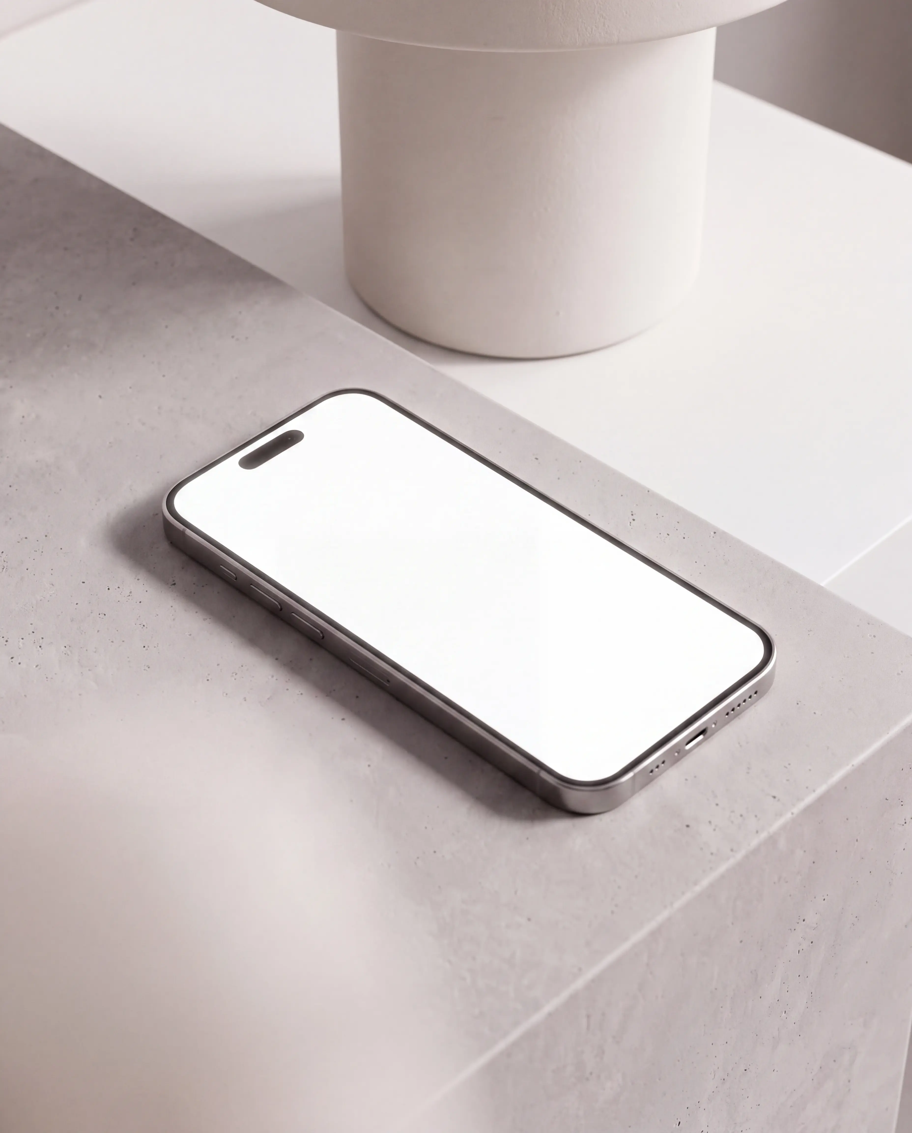 Silver iPhone lying diagonally on a pale stone table beneath a sculptural white side table in a softly lit beige interior, with an editable white screen and Dynamic Island