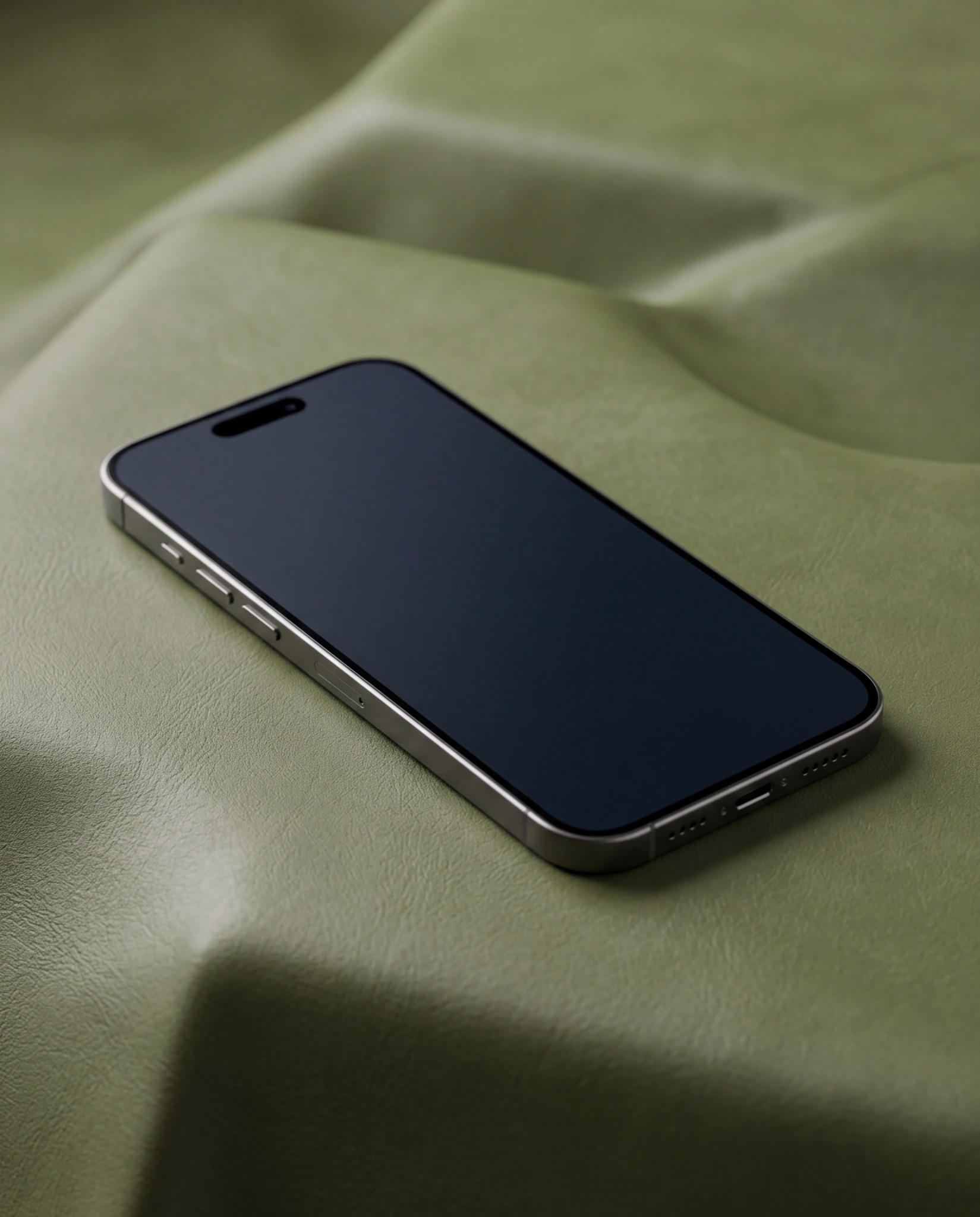 Modern black iPhone lying diagonally on softly folded olive-green leather fabric with an editable vertical screen, visible titanium frame and Dynamic Island